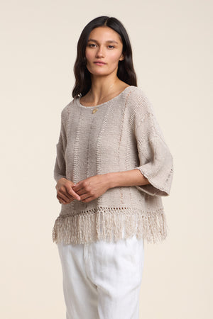 A woman stands against a plain off-white background wearing the KATARINA SWEATER by Velvet by Graham & Spencer—an oversized beige cotton tape sweater with a fringe hem—paired with white pants.
