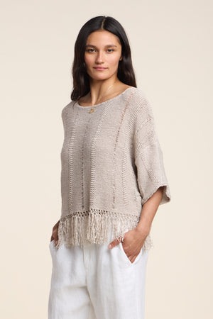 A woman stands with hands in pockets wearing the Velvet by Graham & Spencer KATARINA SWEATER—an oversized, draped cotton tape knit with a fringe hem—paired with white pants against a plain backdrop.