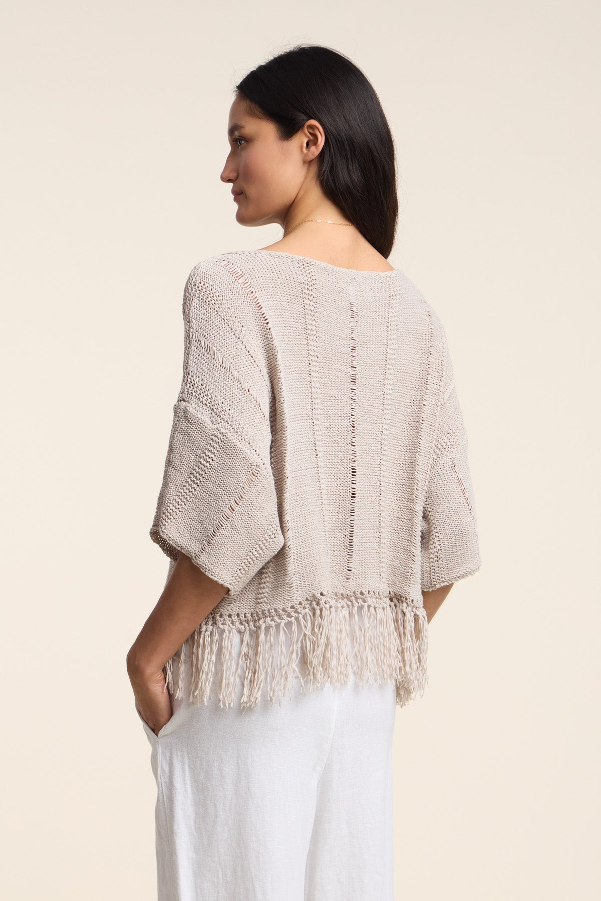 A woman stands sideways against a plain light background, wearing the oversized KATARINA SWEATER by Velvet by Graham & Spencer with fringe hem details, paired with white pants.