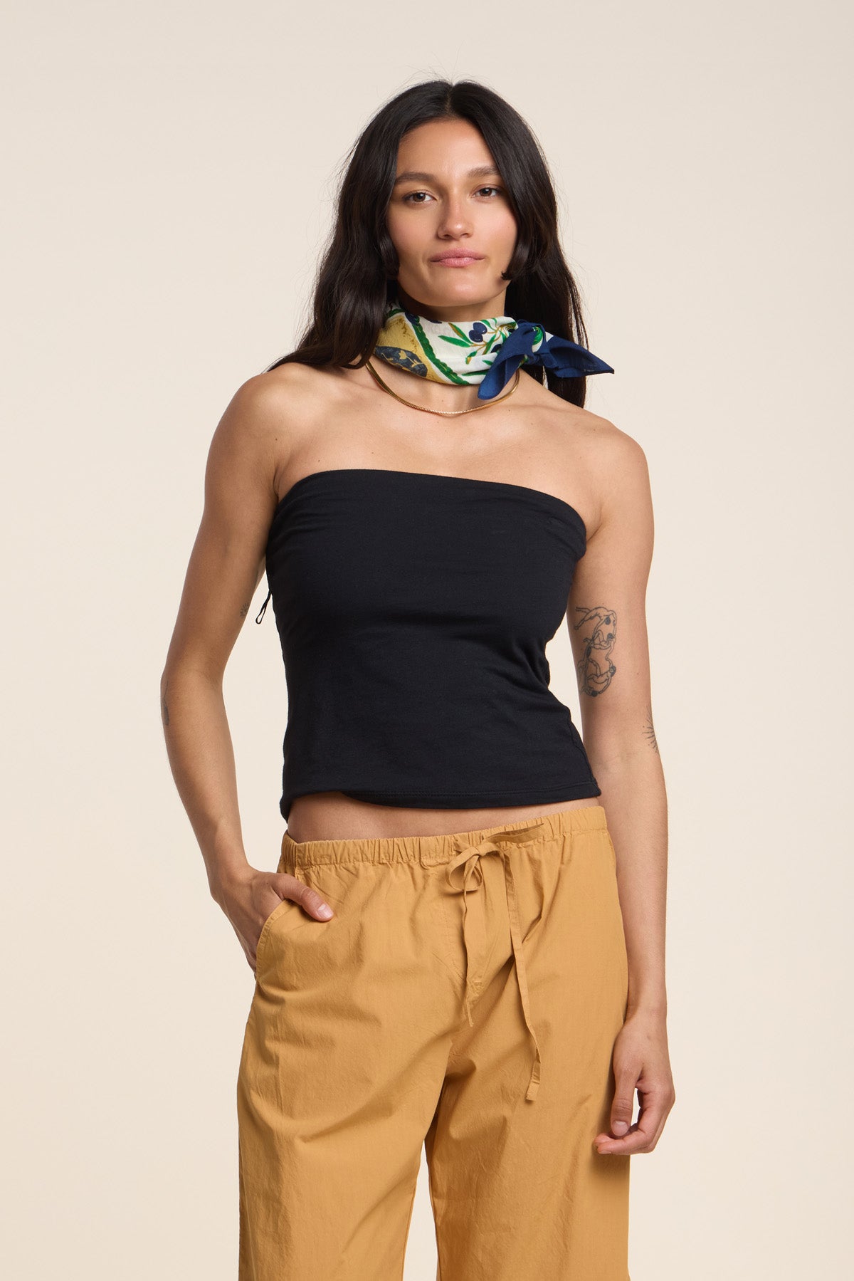 A woman wears the KESS TOP by Velvet by Graham & Spencer—a black, fitted, strapless top—paired with tan drawstring pants and a patterned neck scarf, standing with one hand in her pocket against a plain background.-41371950874817