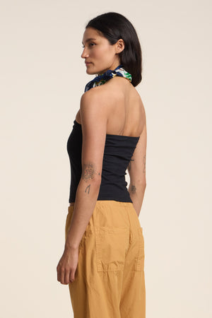 A woman stands in profile wearing the fitted black KESS TOP by Velvet by Graham & Spencer, mustard yellow pants, a neck scarf, and showcasing visible arm tattoos.