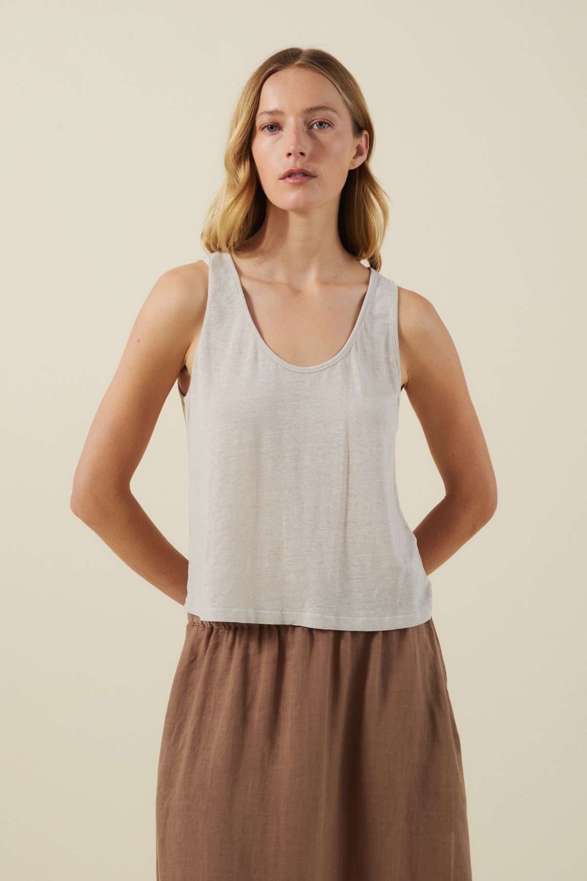 A woman stands against a plain background, wearing a sleeveless KYLIE TANK TOP in light gray by Velvet by Graham & Spencer and a high-waisted brown skirt, with her arms behind her back.