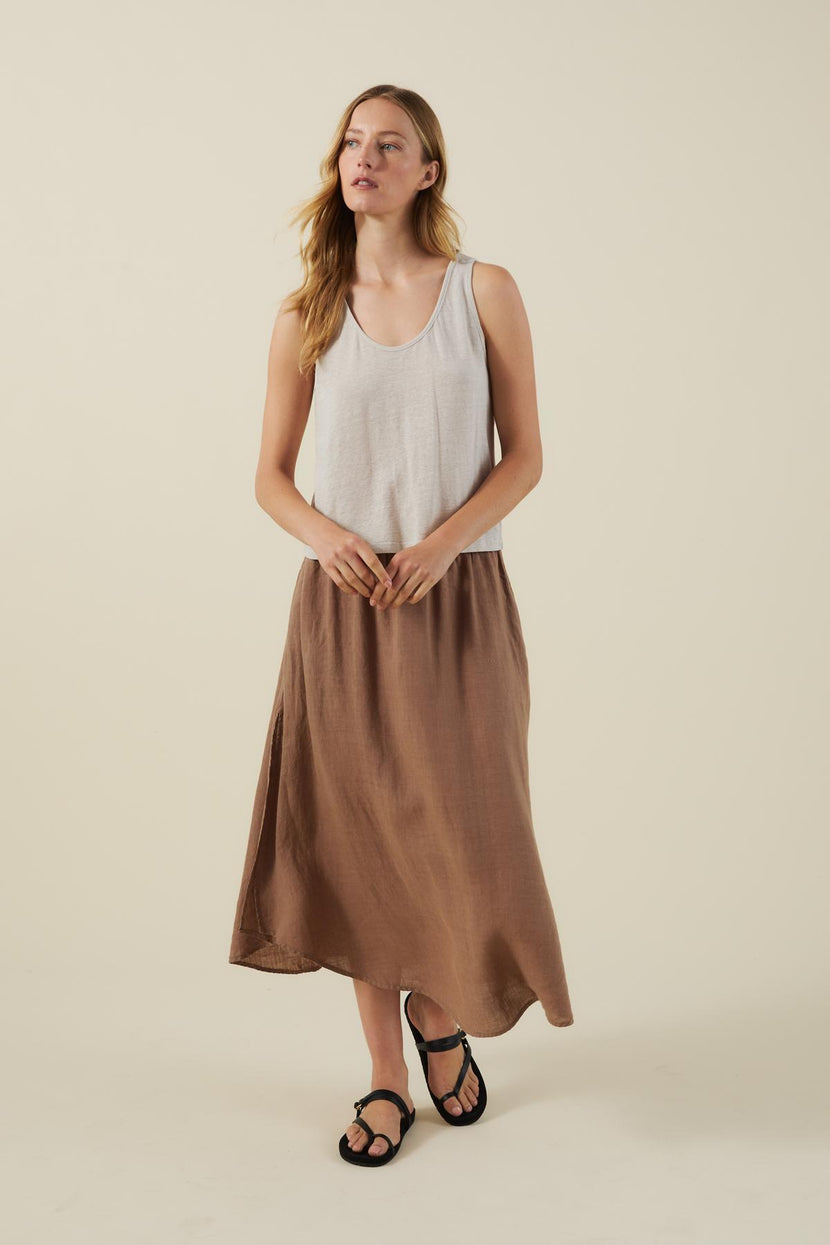 A woman stands against a beige background wearing a white sleeveless top, black sandals, and the VIRGINIA LINEN SKIRT by Velvet by Graham & Spencer—a brown woven linen A-line midi skirt with an elastic waist.