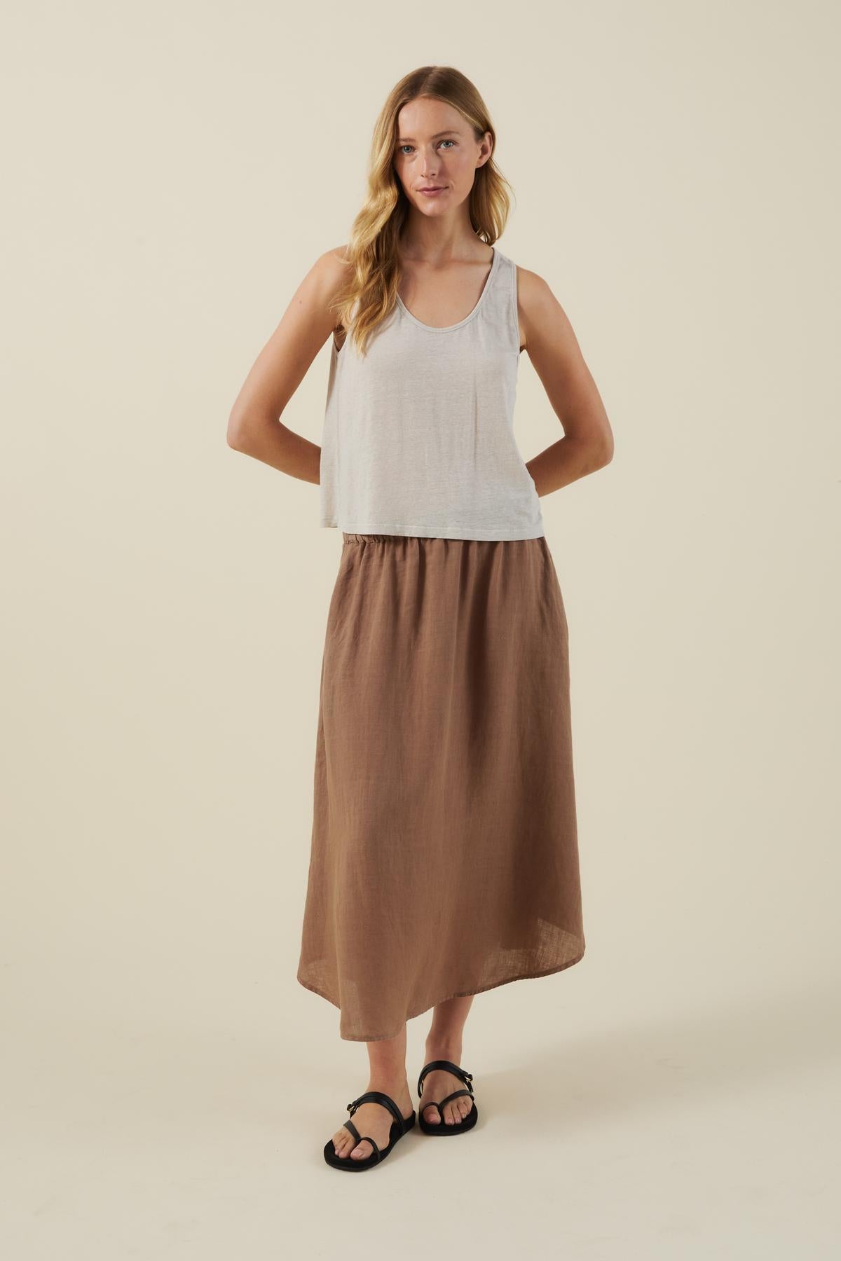 A woman stands against a plain background wearing a white sleeveless top, black sandals, and the VIRGINIA LINEN SKIRT by Velvet by Graham & Spencer—an A-line brown midi skirt with an elastic waist.