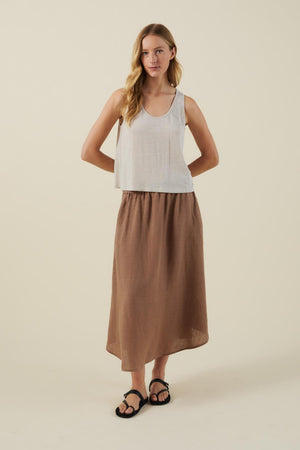 A woman stands against a plain background wearing a white sleeveless top, black sandals, and the VIRGINIA LINEN SKIRT by Velvet by Graham & Spencer—an A-line brown midi skirt with an elastic waist.