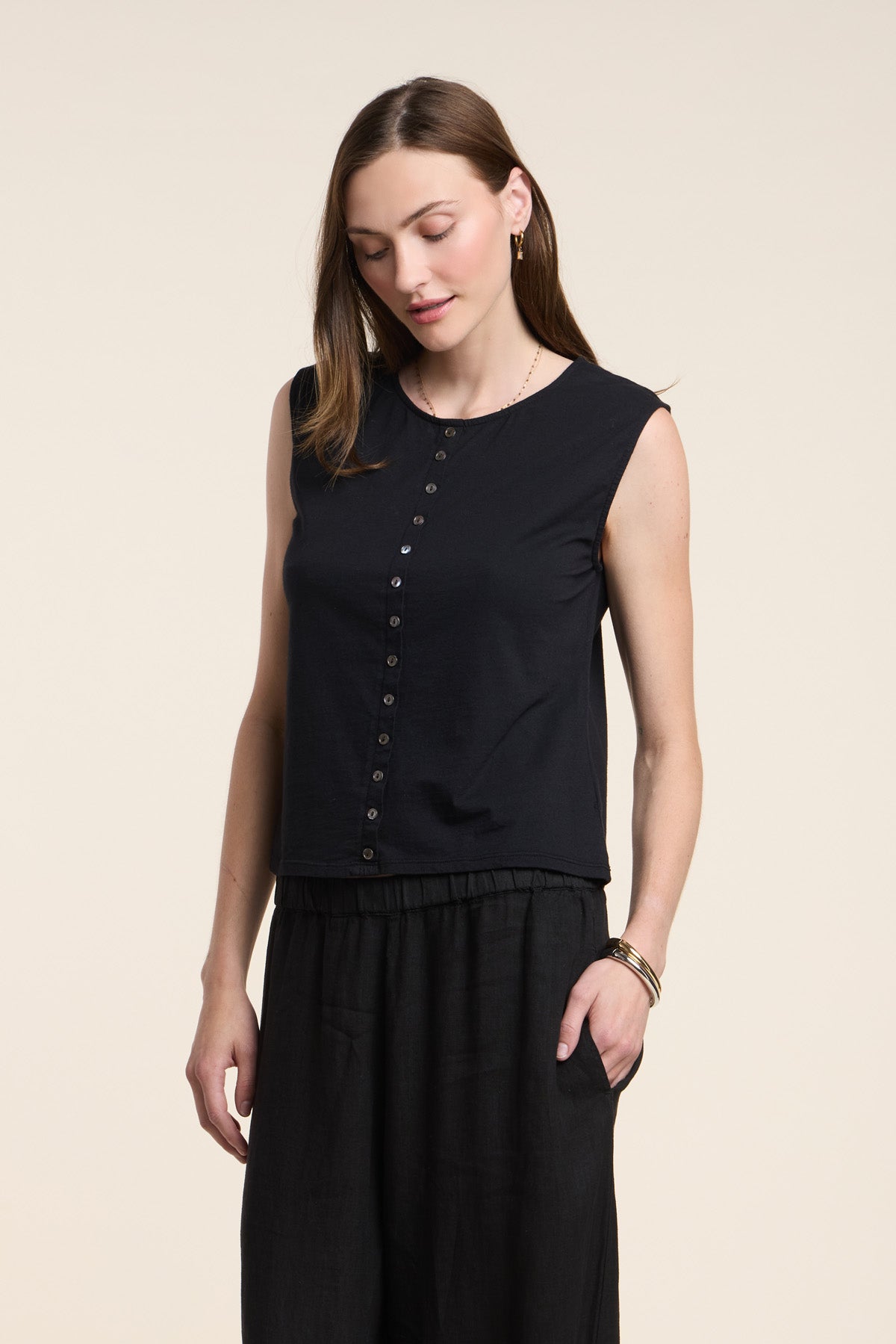 A woman wears the LACY TOP by Velvet by Graham & Spencer—a sleeveless black top with front buttons—paired with black pants, standing with one hand in her pocket against a plain light background.-41371954020545