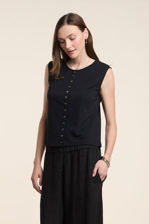 A woman wears the LACY TOP by Velvet by Graham & Spencer—a sleeveless black top with front buttons—paired with black pants, standing with one hand in her pocket against a plain light background.
