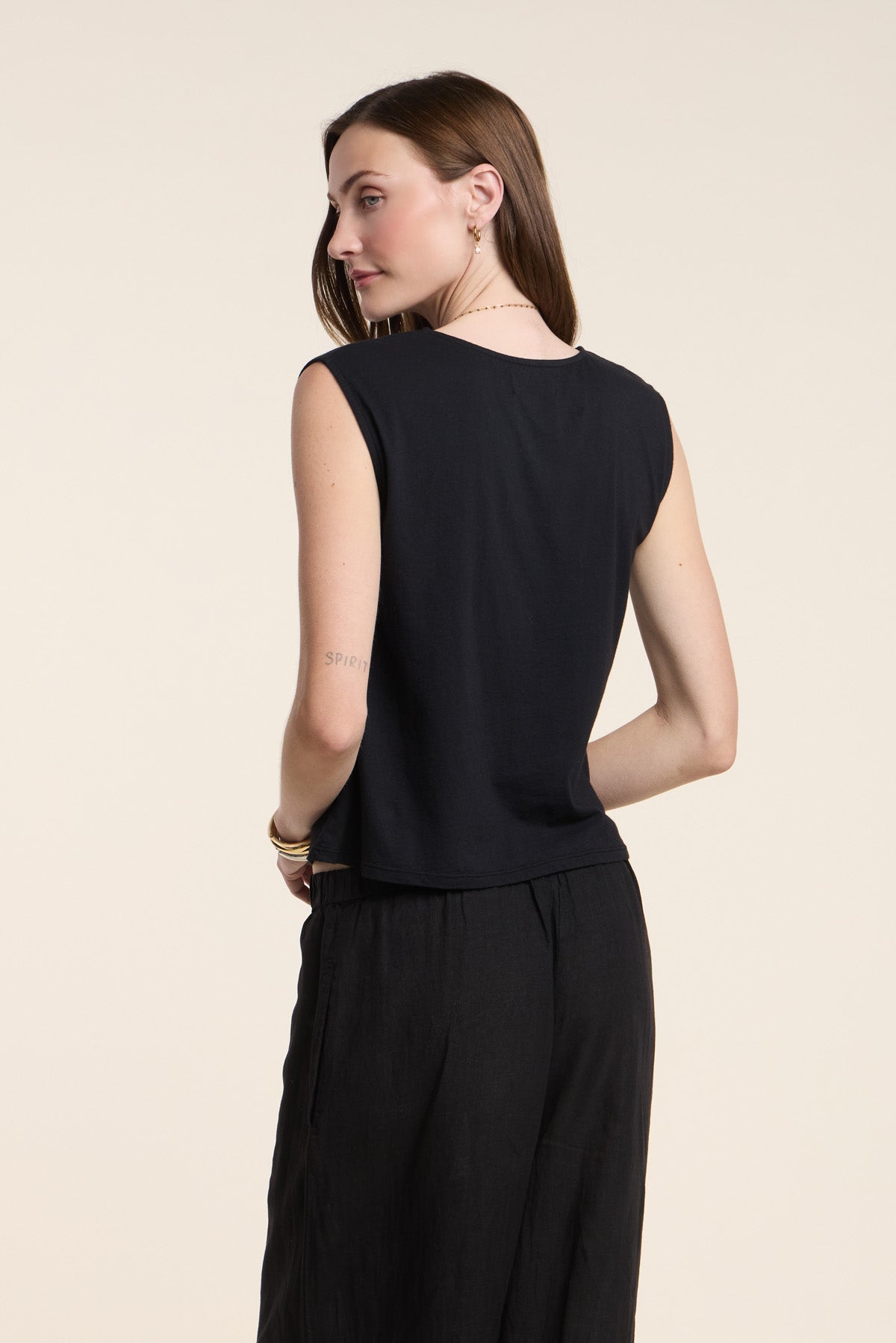 A woman faces away, wearing the sleeveless LACY TOP by Velvet by Graham & Spencer with black pants against a plain light background.