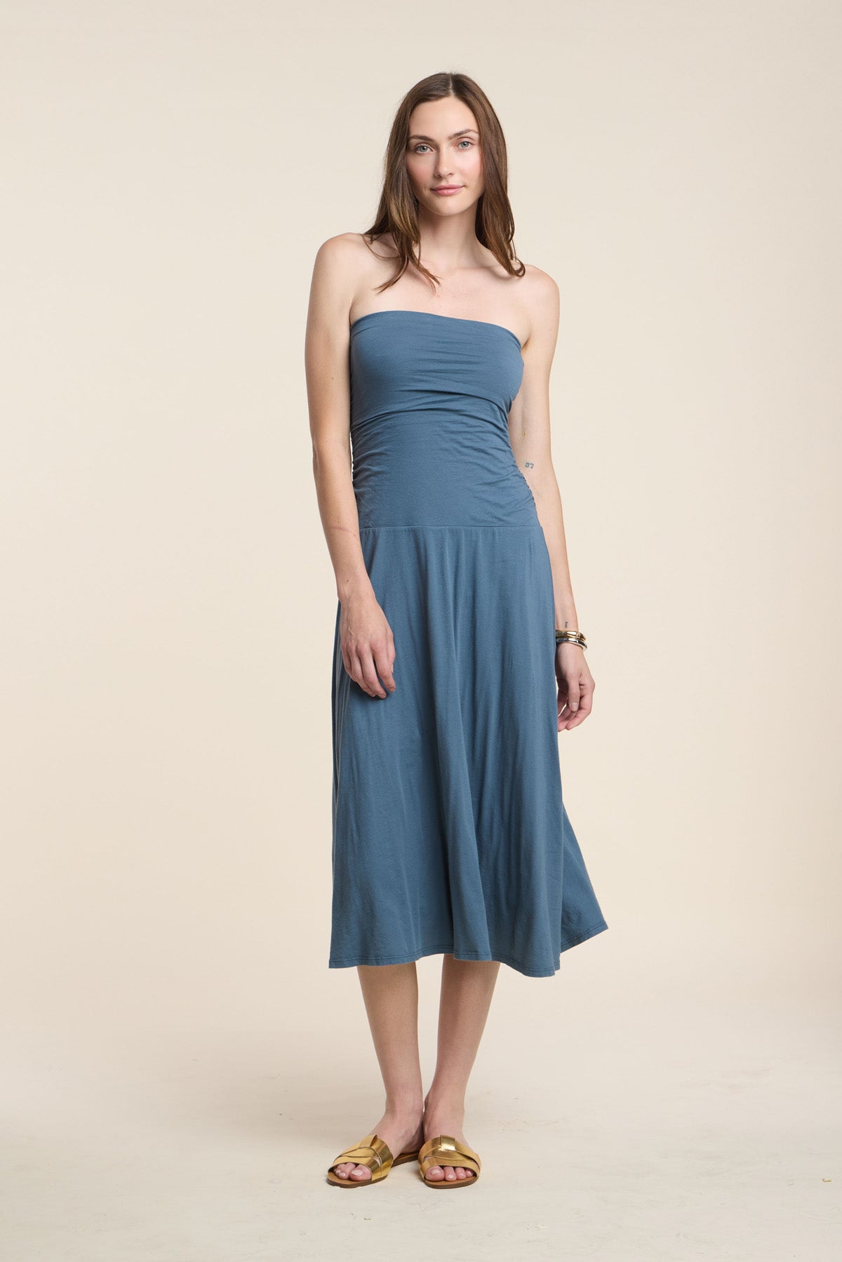 A woman stands against a neutral background wearing the Velvet by Graham & Spencer LAINEY DRESS, a strapless blue midi summer dress, paired with yellow sandals.