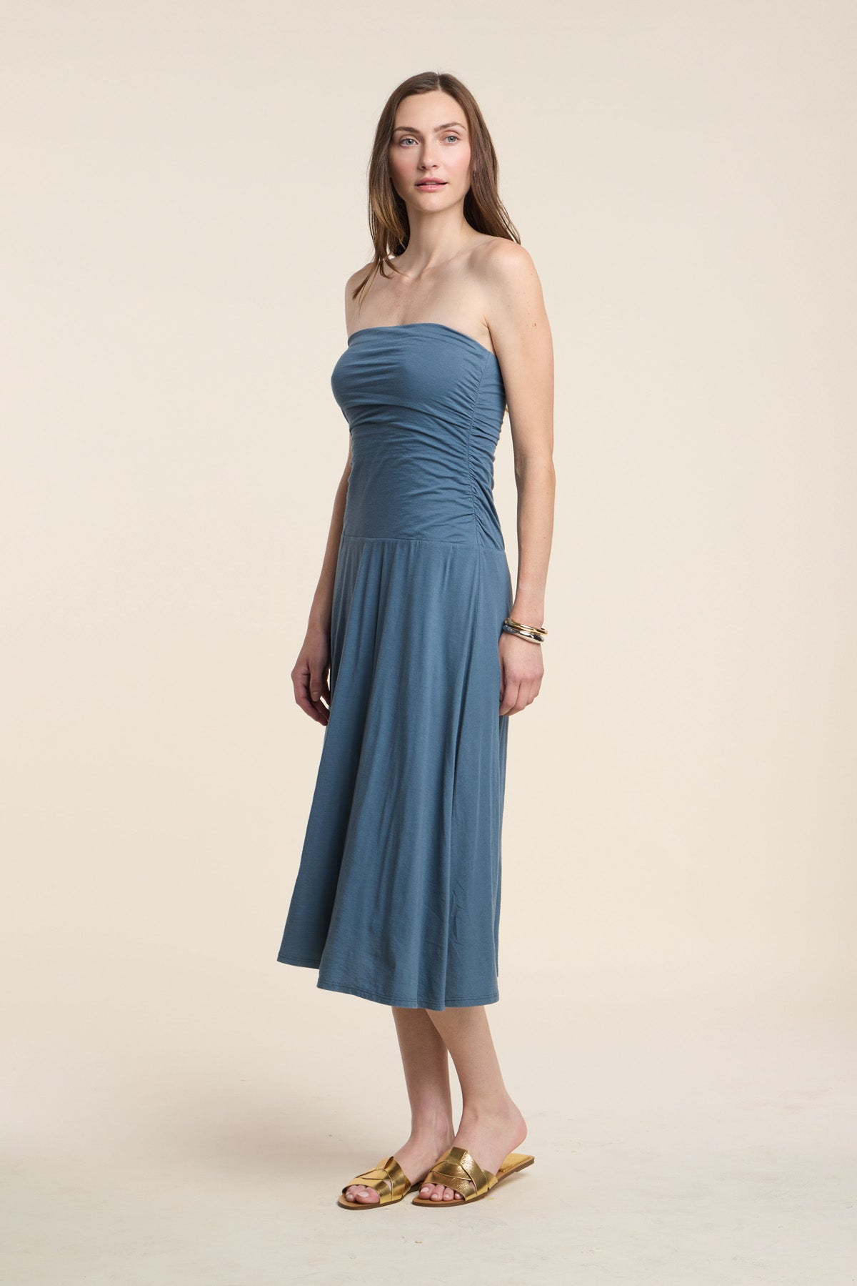 A woman wears the LAINEY DRESS by Velvet by Graham & Spencer, a strapless blue midi summer dress in whisper cotton, paired with gold sandals against a beige background.-41371954380993