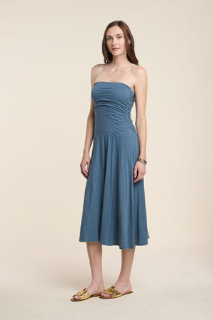 A woman stands against a plain light background wearing the LAINEY DRESS by Velvet by Graham & Spencer, a strapless blue whisper cotton midi, paired with gold sandals; her hair is down and her hands rest relaxed at her sides.