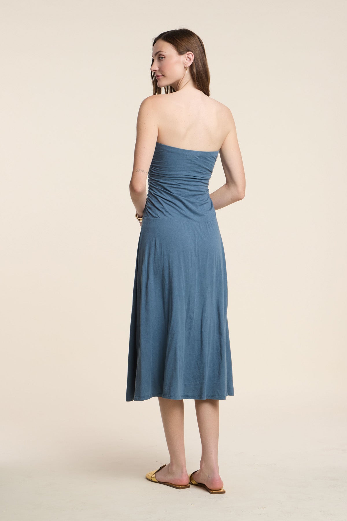 A woman faces away, wearing the Velvet by Graham & Spencer LAINEY DRESS, a blue whisper cotton midi dress, paired with flat sandals against a plain background.