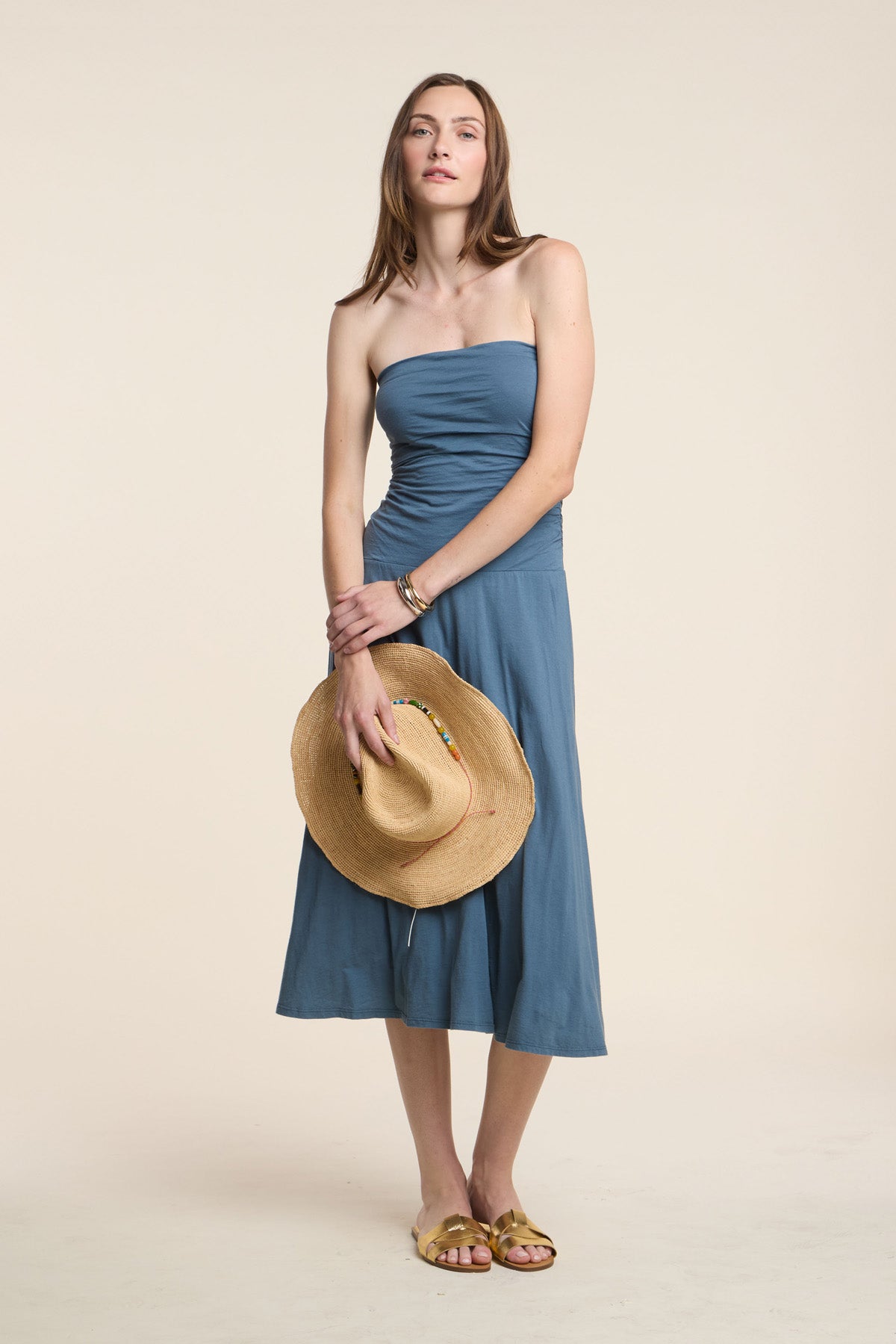 Woman wearing the LAINEY DRESS by Velvet by Graham & Spencer, a strapless blue midi summer dress, holding a straw hat in front of her while standing on a plain light background.-41371954643137