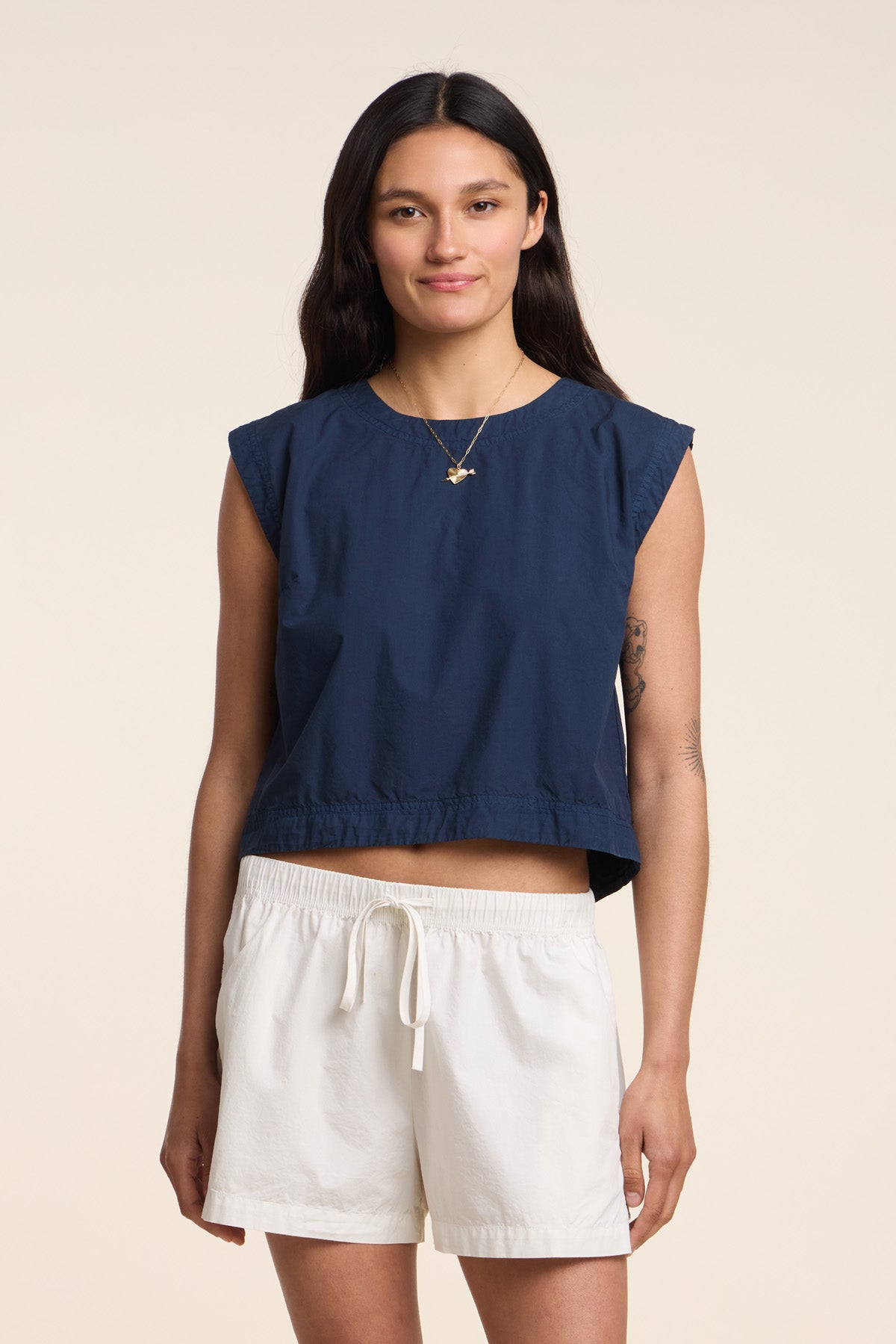 A woman stands facing forward against a plain light background, wearing the LARISSA TOP from Velvet by Graham & Spencer—a navy sleeveless cotton poplin design—paired with white drawstring shorts.