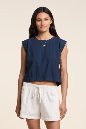 A woman stands facing forward against a plain light background, wearing the LARISSA TOP from Velvet by Graham & Spencer—a navy sleeveless cotton poplin design—paired with white drawstring shorts.