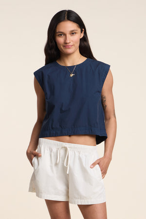 Model wearing the LARISSA TOP by Velvet by Graham & Spencer, a sleeveless cotton poplin top, paired with navy and white drawstring shorts, standing with hands in pockets against a plain light background.