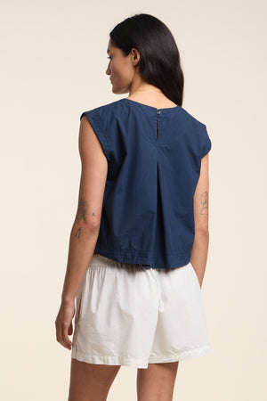 A person stands facing away, wearing the LARISSA TOP by Velvet by Graham & Spencer, a sleeveless navy blue cotton poplin top, paired with white shorts against a plain light-colored background.