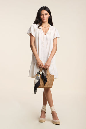 A woman wears the LIVVY LINEN DRESS by Velvet by Graham & Spencer, styled with a straw handbag and beige espadrilles, creating an effortless summer look against a plain background.
