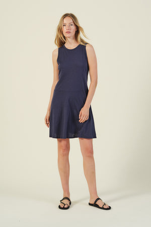A woman poses against a plain light background wearing the LOEN DRESS by Velvet by Graham & Spencer, a sleeveless cotton tank dress in navy blue paired with black sandals—an ideal pick for summer.