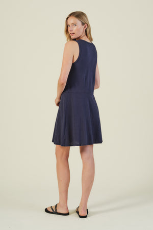 A woman faces slightly away from the camera wearing the LOEN DRESS by Velvet by Graham & Spencer—a sleeveless cotton tank dress—paired with black sandals, set against a simple light background.
