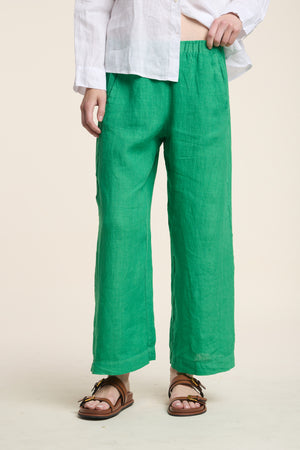 A person wears the Velvet by Graham & Spencer LOLA LINEN PANT in bright green, paired with a white long-sleeve shirt and brown sandals, standing against a neutral background.