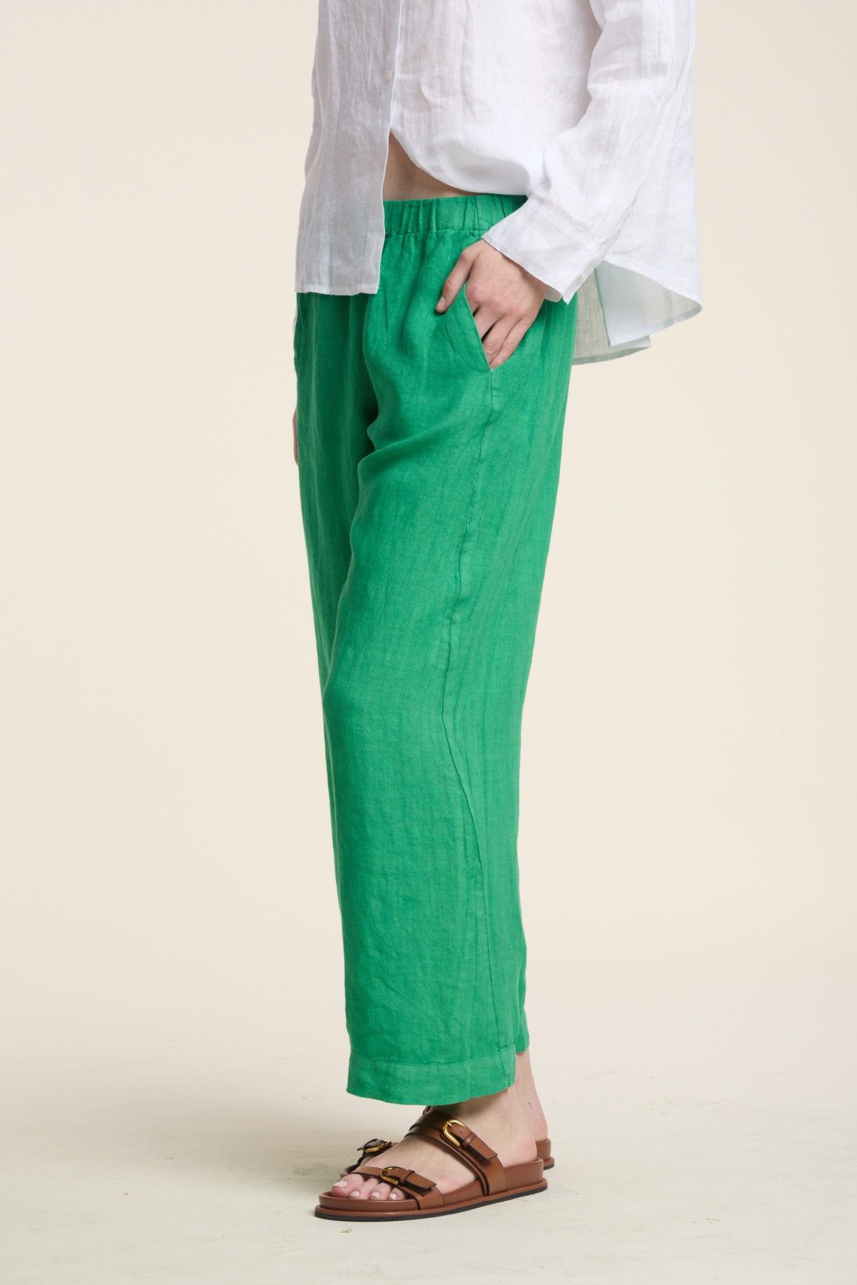 A person stands against a plain background with one hand in their pocket, wearing the LOLA LINEN PANT by Velvet by Graham & Spencer in green, paired with a white long-sleeve shirt and brown sandals.-41527074357441