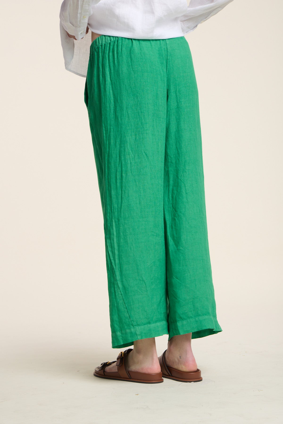 Model wears the LOLA LINEN PANT by Velvet by Graham & Spencer, styled in loose, wide-leg green linen with brown sandals, standing on a light background facing away from the camera.-41527074390209