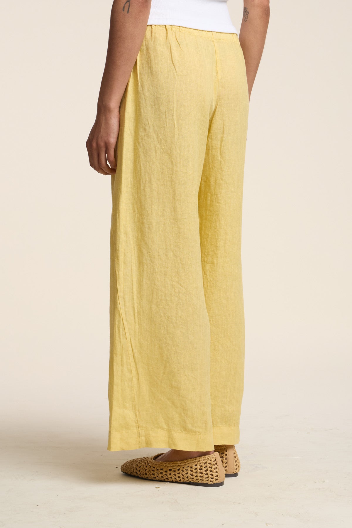 Wearing the Velvet by Graham & Spencer LOLA LINEN PANT—loose-fitting yellow linen pants with a relaxed straight leg—a person pairs them with a white top and woven tan shoes, photographed from behind against a neutral background.