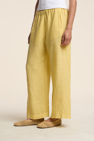 A person wearing the LOLA LINEN PANT in yellow by Velvet by Graham & Spencer, paired with a white top and woven beige flats, stands against a plain light background.
