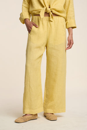 A person wears the LOLA LINEN PANT by Velvet by Graham & Spencer in yellow, paired with a matching tied-waist shirt and woven beige flats, standing against a plain background.