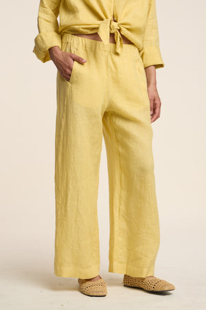 Model wears the LOLA LINEN PANT by Velvet by Graham & Spencer with a matching long-sleeve shirt tied at the waist and woven beige slip-on shoes, standing against a plain background.