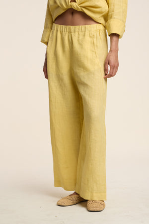 Wearing the Velvet by Graham & Spencer LOLA LINEN PANT—loose, wide-leg, yellow linen pull-ons—paired with a matching top and beige woven slip-ons, standing against a plain background.
