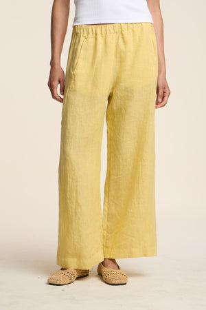 A person is wearing the LOLA LINEN PANT by Velvet by Graham & Spencer—loose-fitting, yellow linen pants with a relaxed straight leg—paired with a white top and woven beige shoes, standing against a plain background.