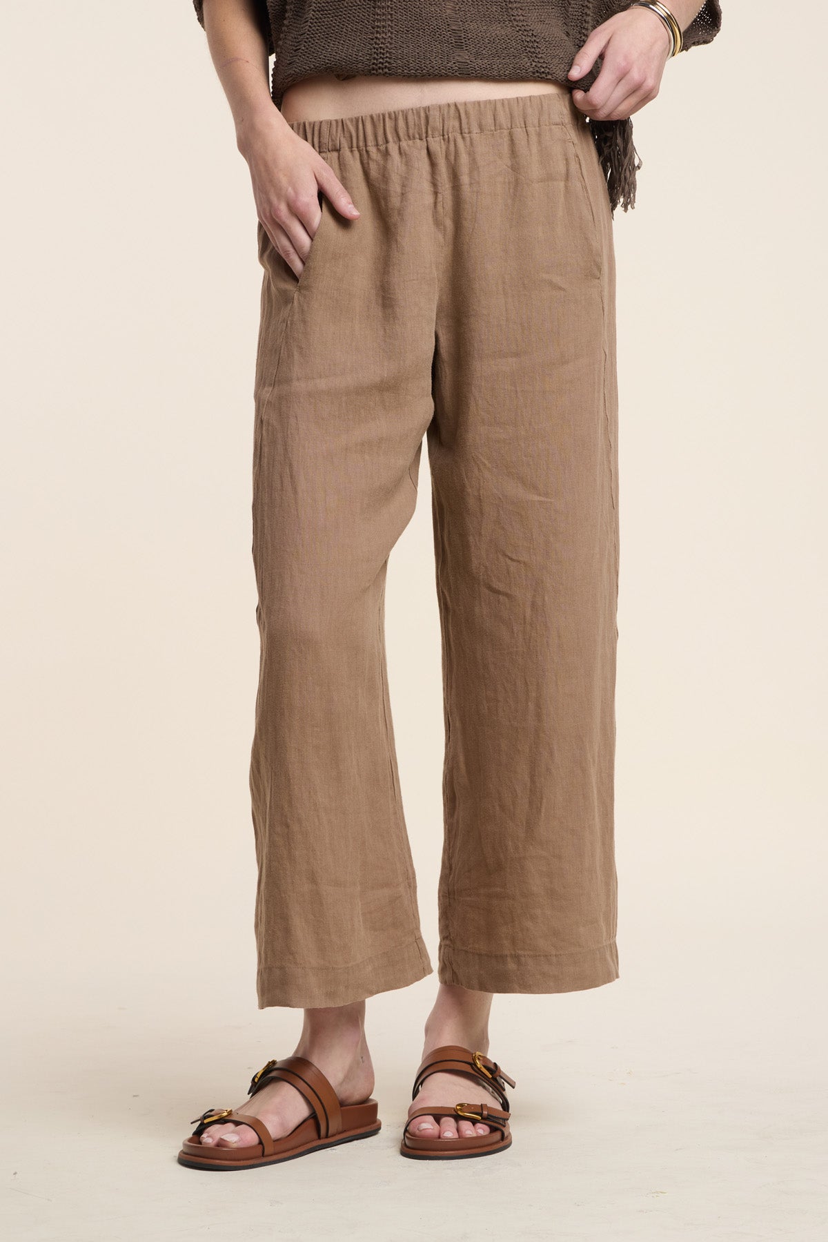 Model wears the LOLA LINEN PANT by Velvet by Graham & Spencer—cropped, lightweight linen pants—paired with brown sandals, posing with one hand in a pocket against a plain background.-41527067836609