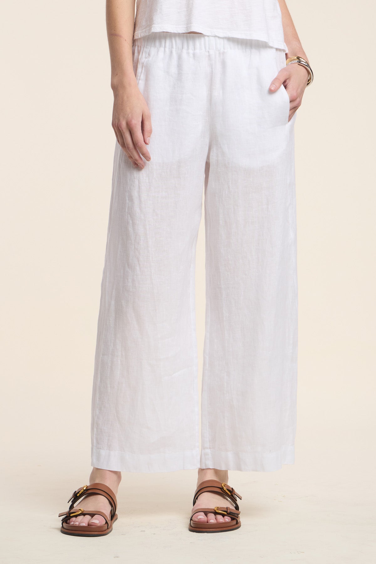 A person is wearing the LOLA LINEN PANT by Velvet by Graham & Spencer—a wide-leg, loose-fitting white linen pant—paired with a white top, brown sandals, and a bracelet, standing against a plain light background.-41803019813057