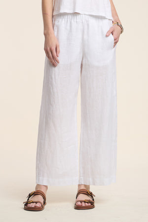 A person is wearing the LOLA LINEN PANT by Velvet by Graham & Spencer—a wide-leg, loose-fitting white linen pant—paired with a white top, brown sandals, and a bracelet, standing against a plain light background.