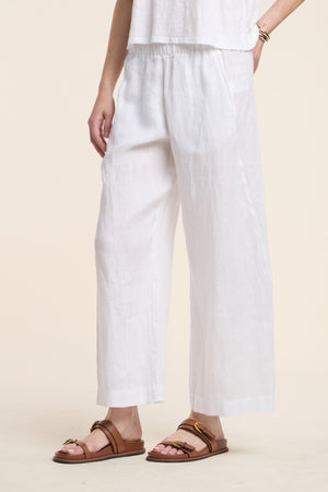 A person wearing the LOLA LINEN PANT by Velvet by Graham & Spencer in white, paired with a white top and brown sandals, stands against a neutral background. Only the lower torso and legs are visible.