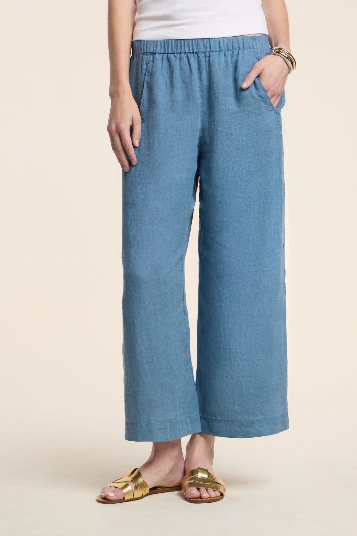 Wearing the LOLA LINEN PANT by Velvet by Graham & Spencer, paired with a white sleeveless top, gold bracelets, and gold sandals; standing with one hand in the pocket.