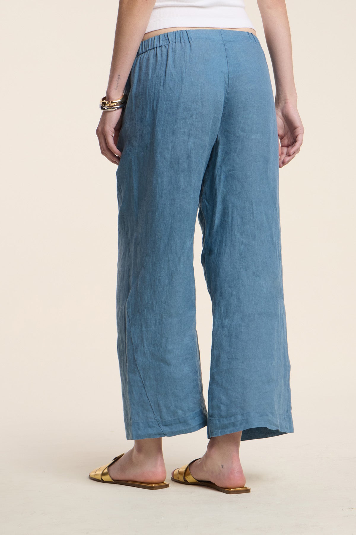 Model wears the Velvet by Graham & Spencer LOLA LINEN PANT in light blue—lightweight linen with an elastic waistband and wide leg—paired with a white top, yellow sandals, and a bracelet, back turned to camera against a neutral backdrop.-41385454928065