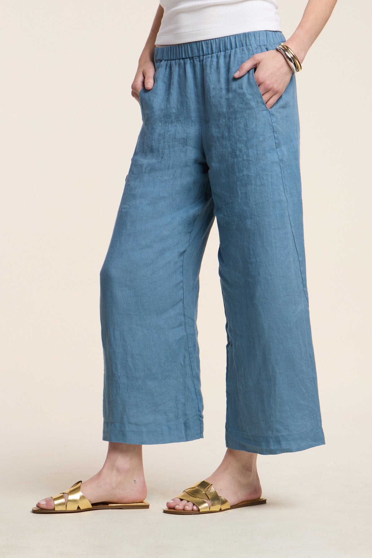 A person wearing Velvet by Graham & Spencer’s LOLA LINEN PANT with an elastic waistband, a white top, gold slide sandals, and a bracelet stands with hands in pockets.-41385454895297