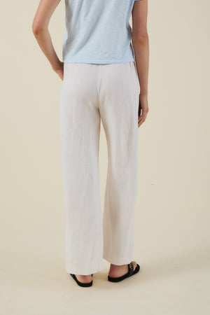A person stands with their back to the camera against a plain beige background, wearing a light blue short-sleeve top, black sandals, and the LOLA LINEN PANT by Velvet by Graham & Spencer in white with a wide-leg silhouette.