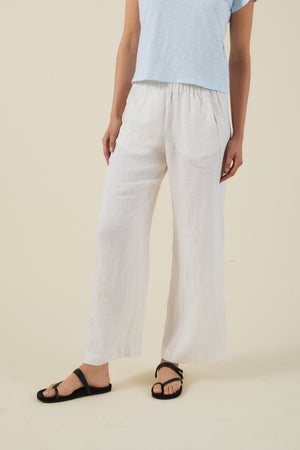 A person wearing a light blue short-sleeve top, black sandals, and the LOLA LINEN PANT by Velvet by Graham & Spencer—white, wide-leg lightweight linen pants—stands against a plain beige background.