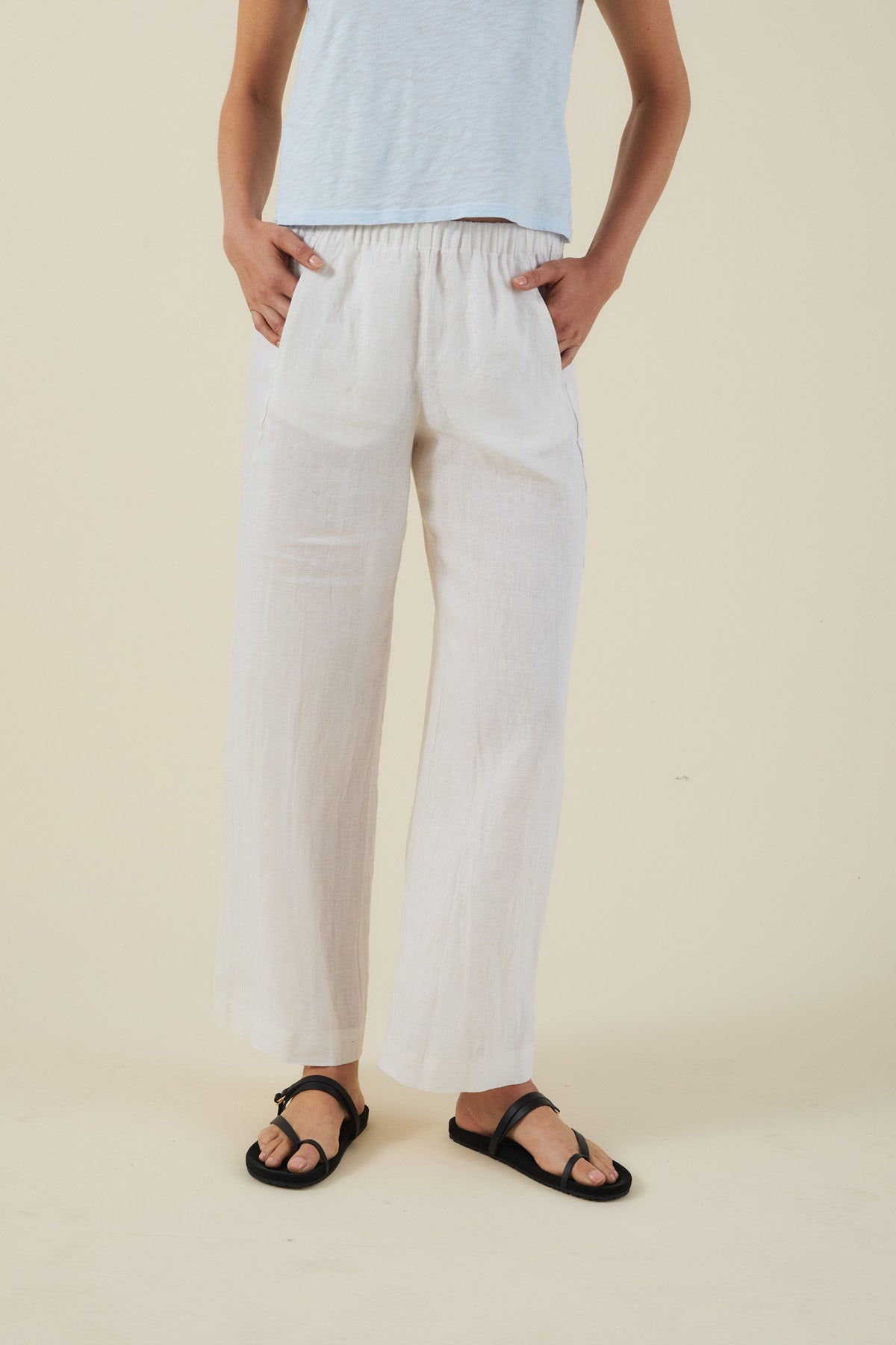 A person is wearing a light blue top, Velvet by Graham & Spencer’s LOLA LINEN PANT in white, and black sandals, standing against a plain beige background.-41779861356737