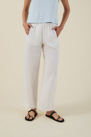 A person is wearing a light blue top, Velvet by Graham & Spencer’s LOLA LINEN PANT in white, and black sandals, standing against a plain beige background.