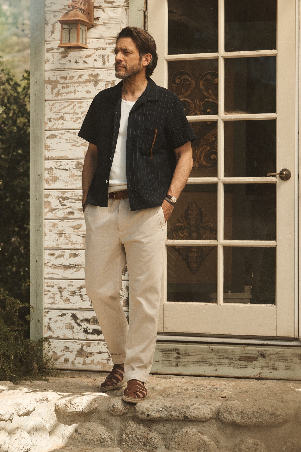 A man stands on stone steps outside a house, wearing the GUNNER SHIRT by Velvet by Graham & Spencer over a white tee, beige pants, and brown sandals, hands in pockets, looking to the side.