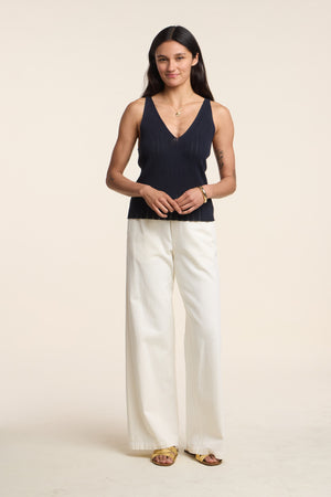 A woman stands against a plain background wearing a navy sleeveless V-neck top, the LOUISE DENIM PANT by Velvet by Graham & Spencer with a relaxed wide-leg fit, yellow sandals, and simple jewelry.