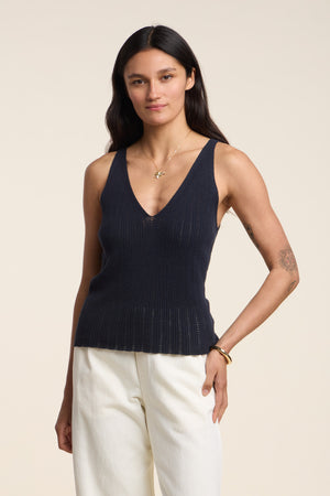 A woman with long dark hair wears the LORENA TANK TOP by Velvet by Graham & Spencer in navy, paired with white pants, ideal for warm-weather layering, standing against a plain light background.