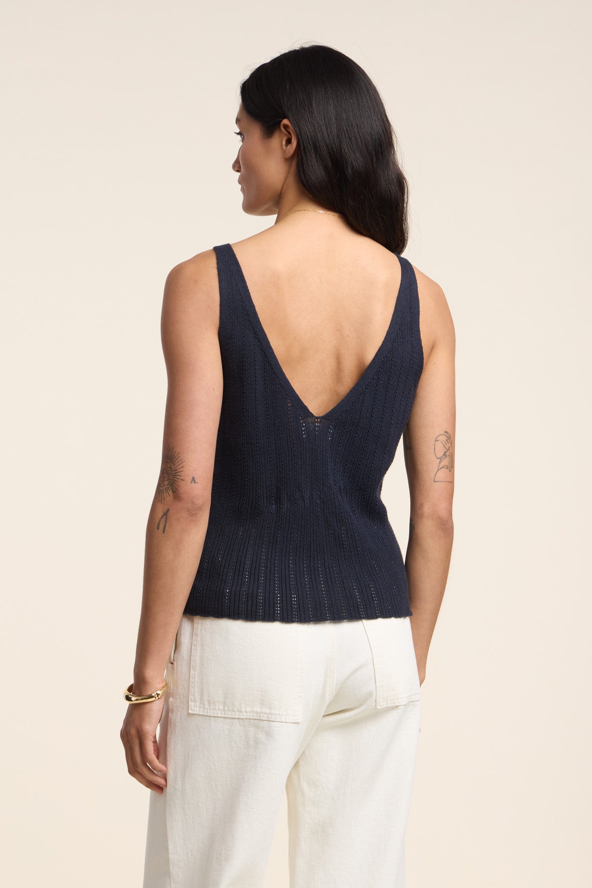 A woman with long dark hair faces away, wearing the LORENA TANK TOP by Velvet by Graham & Spencer—an organic cotton-linen blend sleeveless navy top with a deep V-back—paired with white pants, perfect for warm-weather layering.-41527093264577
