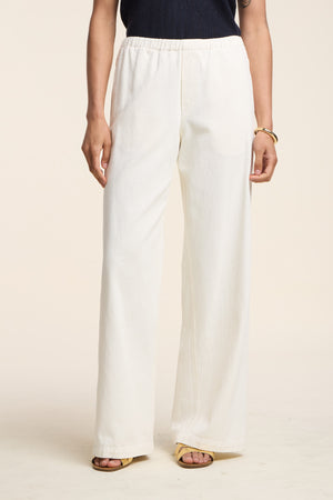 A person in Velvet by Graham & Spencer’s LOUISE DENIM PANT, a black sleeveless top, yellow sandals, and a bracelet stands against a plain background.
