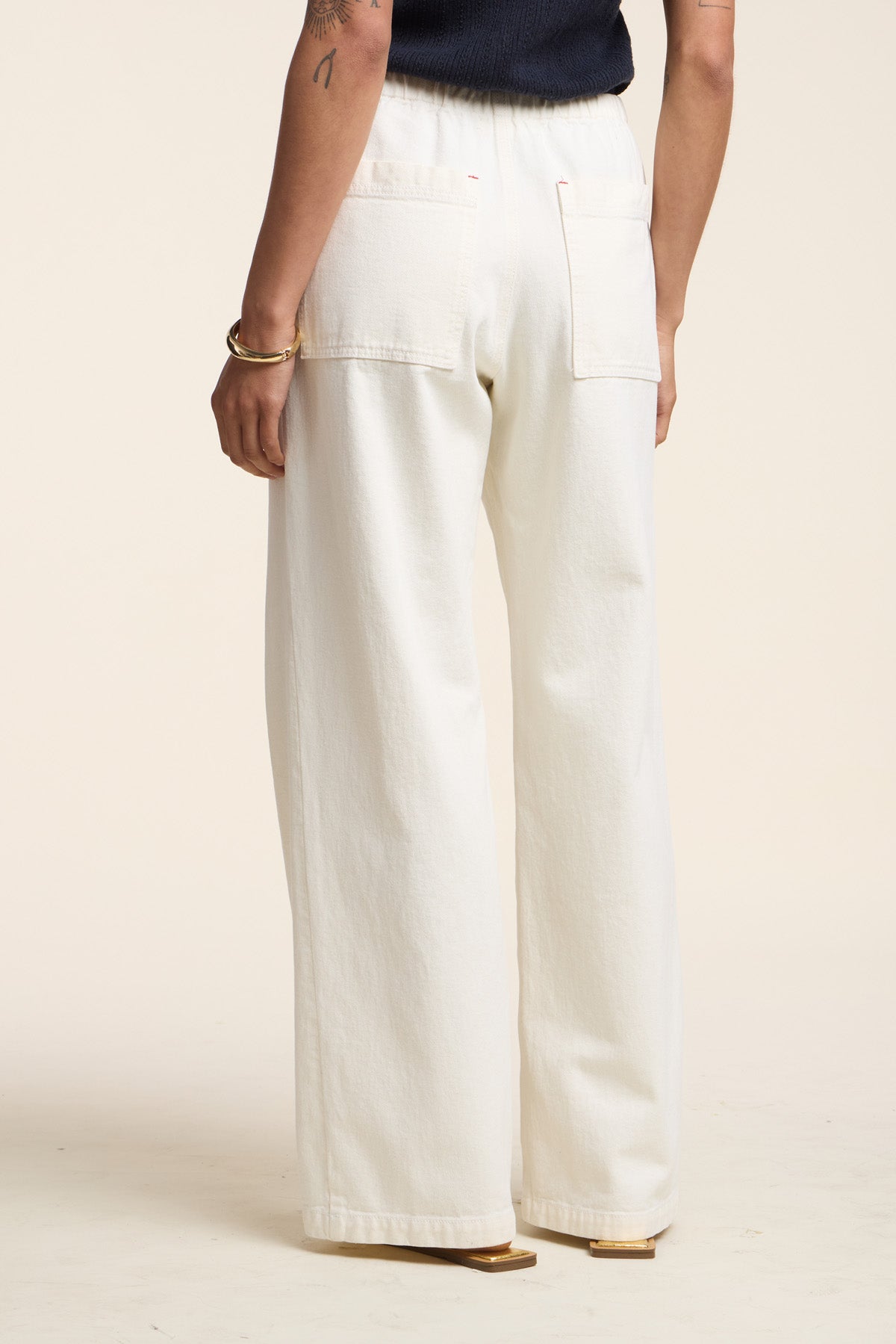 A person stands with their back to the camera, wearing Velvet by Graham & Spencer's LOUISE DENIM PANT—high-waisted, wide-leg cotton twill with large back pockets—and a sleeveless black top for a relaxed look.-41527096410305